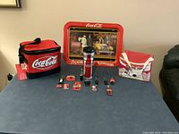Full view of all items in the lot on a table: two cooler bags, one metal TV tray, thermo glass, and 12 Coca-Cola magnets.