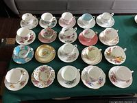 Top-down view of twenty assorted vintage tea cup and saucer sets displayed on dark cloth showing variety of floral patterns and gold trimming.