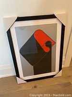 Front view of the framed abstract print showing the geometric artwork in black, gray, and orange inside a black frame with white mat and cardboard protective corners.