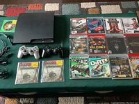Black Sony PlayStation 3 console with 2 controllers (silver and black), power cable, AV cable, USB cable, and scattered game cases on green surface.