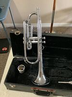 Front view of silver trumpet inside open black hard shell case on beige surface showing valve oil container and mouthpiece.