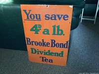 Front view of the vintage porcelain sign showing bold orange background with blue and green text advertising Brooke Bond Dividend Tea and savings of 4d per pound.