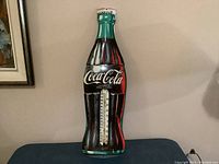 Front view of vintage Coca Cola metal thermometer sign showing Coca Cola logo and space for thermometer in center.