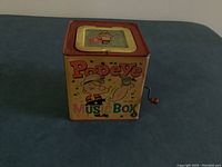 Photo of the front of the vintage metal square music box with Popeye graphics and red crank handle on the side.