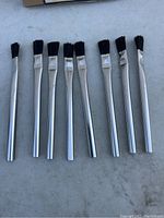 Eight individual acid brushes with black horsehair bristles and shiny silver metal handles arranged in a row on a white background.