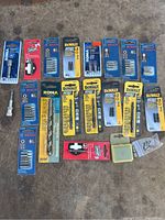 Photo showing assorted retail-packaged drill and driver bits from Dewalt, Bosch, Rona, and Vermont American brands arranged together.