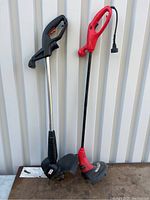 Two electric weed whackers standing upright, one black with silver shaft and the other red and black, both with cords.