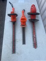 Three orange electric hedge cutters displayed side by side on a white table surface