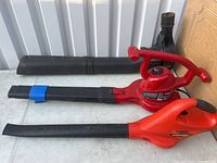 Three components on a light-colored table: one Black & Decker leaf blower, one red Toro leaf blower, and a long black blower hose attachment.