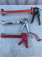 Photo of three caulking guns showing red and chrome metal designs on a white surface.