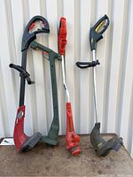 Four electric weed whackers standing upright by a wall showing different colors and design features.