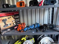 Metal shelving with four power tools displayed: boxed cordless drill/driver, two corded grinders, and one corded drill. Some tools have visible power cords coiled.
