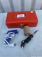 Red metal toolbox with soldering iron, glue gun, and spool of solder wire arranged on tabletop.
