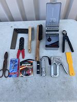 Image showing assortment of tools including wire stripper, stapler, wire brushes, scissors, flashlight, and scraper on a white surface