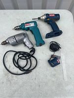 Three drills displayed on a table: metallic corded drill with power cable, teal Makita cordless drill, and blue Ryobi cordless drill with charger.