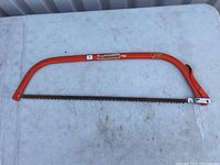 Photo showing the entire Mastercraft bow saw on a gray surface, displaying the orange metal frame with the saw blade fixed between the ends.