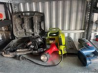 Shelving area holding all the tools showing the black drill case, battery charger, Ryobi green electric chainsaw, red flashlight, and Ryobi blue reciprocating saw.