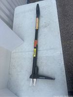 Image of a black and orange Fiskars extendable weed ejection tool with soft grip handle and metal fork at the base, laying on a white surface.