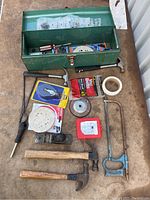 Tools and accessories laid out next to green metal toolbox, showing hammers, hacksaw frame, sanding discs, hand plane, tape and metal spool.