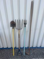 Photo showing four garden tools: shovel, pitchfork, small cultivator fork, and hoe, against a white corrugated background.