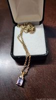 Photo showing gold-toned chain with rectangular faceted purple pendant on white cushion in black box.