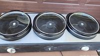 Photo of the electric slow cooker with three round pots and glass lids placed on top, showing the front control knobs.