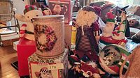 Wide view of Christmas decoration lot showing snowman plush, metal tin, Santa figures, and multiple ornaments on table.