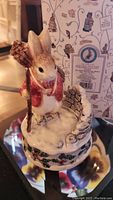Photo showing Peter Rabbit ceramic music box holding a broom, standing on snowy base with holly design and stone wall detail, with original box in background.