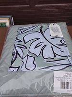 Showing folded picnic blanket inside clear plastic packaging with visible sketched palm leaf design and brand tag.