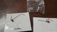 Two silver-tone necklaces on branded backing card, one with small red gemstone pendant, other with blue pendant, plus pair of blue gemstone earrings sealed in plastic bag