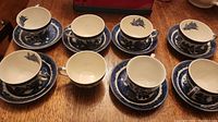Eight cups and seven matching saucers arranged on a wooden surface, showing blue Willow pattern decoration on white ceramic.
