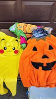 Photo of three kids costumes arranged together: Pokemon Pikachu yellow costume, orange pumpkin baby costume from Pottery Barn, and a folded Batman small costume with cape.