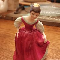 Porcelain figurine showing front and side view of the Royal Doulton 'Fair Maiden' figure in a red dress with white sleeves and yellow bow in hair.