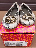 Front view of pair of Tory Burch snake print flats with gold-tone logo on toe sitting on original branded box.