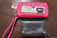 Photo of two wallets: pink Mundi Under Cover wallet with wrist strap and tags, and black Roots wallet with white stitching.
