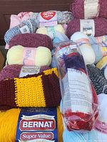 Lot of assorted yarn skeins including Bernat Super Value yellow yarn, burgundy textured yarns, and baby yarns in light yellow and cream with a crocheted two-tone scarf.