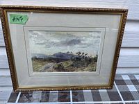 Framed watercolor painting showing a landscape with a road, mountains, and cloudy sky. The frame is gilded wood with decorative edges and a matting around the painting.
