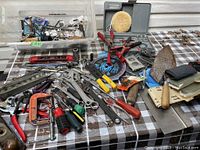 Wide shot showing assortment of hand tools including wrenches, clamps, pliers, screwdrivers, and miscellaneous hardware spread out on checkered fabric.
