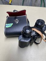 Black 8x30 Comet binoculars resting on top of black leather carrying case with snap closure flap open showing red interior.