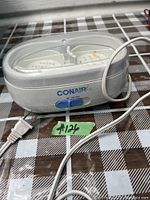 White Conair electric jewelry cleaner with transparent lid and power cord on checkered surface.