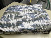 Photo showing the folded Eddie Bauer blanket with a nature pattern of trees in gray, white, and green on the exterior.