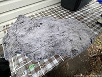 Soft grey fake fur rug resting on a plaid surface, shows overall texture and irregular edges.