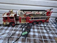 Photo of the left side of a large red and gold Metro fire department toy fire truck with attached remote control and connected wire, showing the truck length, wheels, and ladder details.