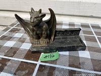 Photo of a metal-like gargoyle figurine with a muscular physique, wings extended behind, perched on a rectangular base with decorative details.