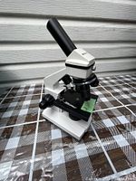 White microscope angled showing eyepiece, stage, and adjustment knobs on a plaid surface with siding background.