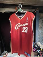 Front full view of red Cleveland Cavaliers LeBron James 23 basketball jersey hanging on a hanger showing team name, number and overall condition.