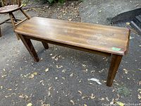 Wooden bench placed outdoors on ground covered with dry leaves and dirt, showing the full side and top surfaces and legs of the bench.