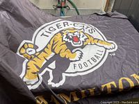 Close-up of the Hamilton Tiger-Cats football flag showing the leaping tiger logo and the word 'HAMILTON' in gold letters on a dark fabric background.