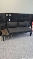 Black metal framed bench with wooden side table on the left and dark gray cushions, photographed from front and slightly side angles.