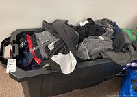 Photo showing the large black plastic tote filled with assorted men's clothing including pants, shorts, and shirts in various dark colors.